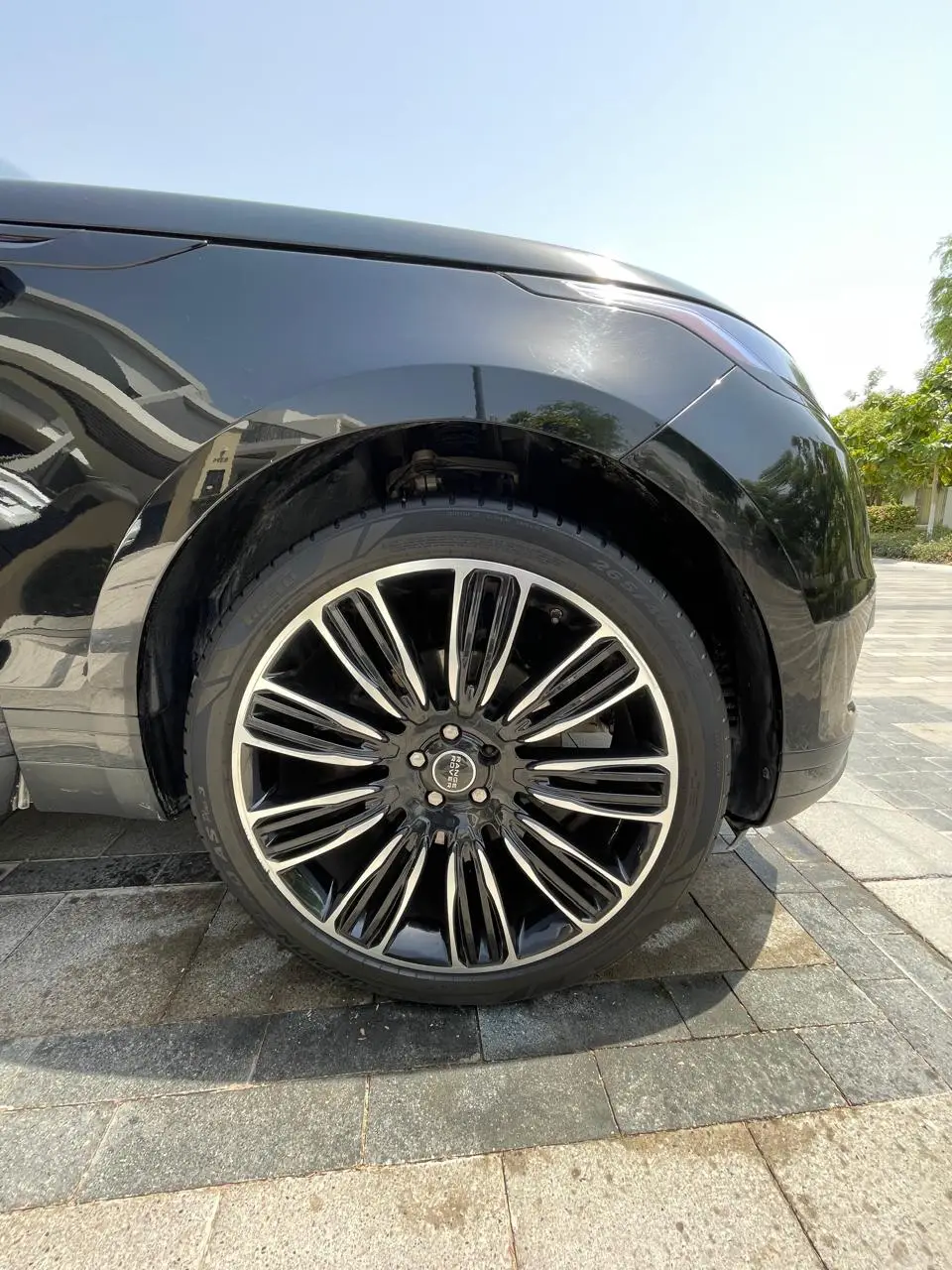 Close-up of a black Range Rover’s front wheel, tire, and reflective front fender, parked on a paved driveway in sunlight.