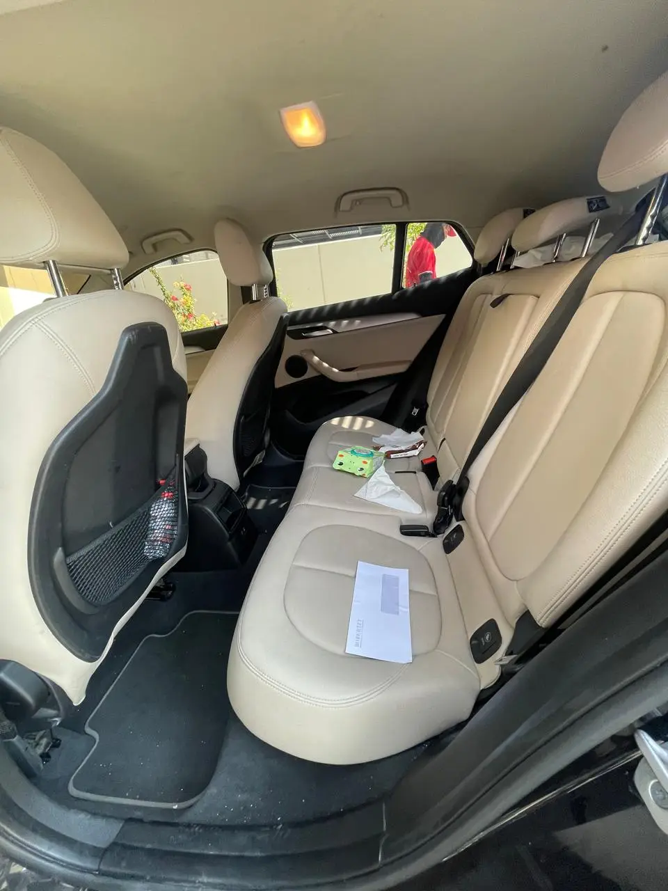 View through an open rear car door showing beige leather seats, black floor mats with scattered debris, and tissues, a packet, and an envelope on the back seat.