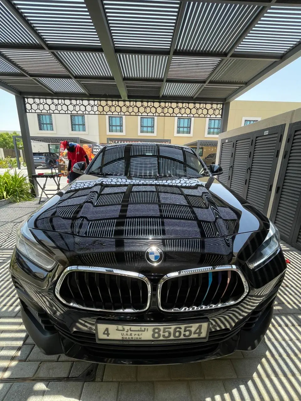 Front view of a glossy black BMW under a slatted outdoor canopy, with striped reflections across the hood and a worker in red beside the car.