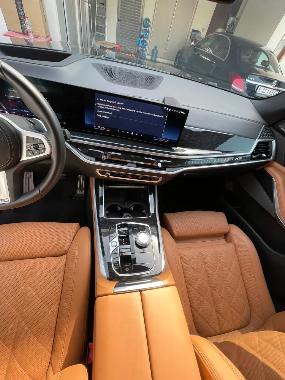 BMW X5 interior with tan leather seats, a black dashboard, and a center console. The dashboard screen displays a key-not-recognized warning, and another car is visible in the garage ahead.