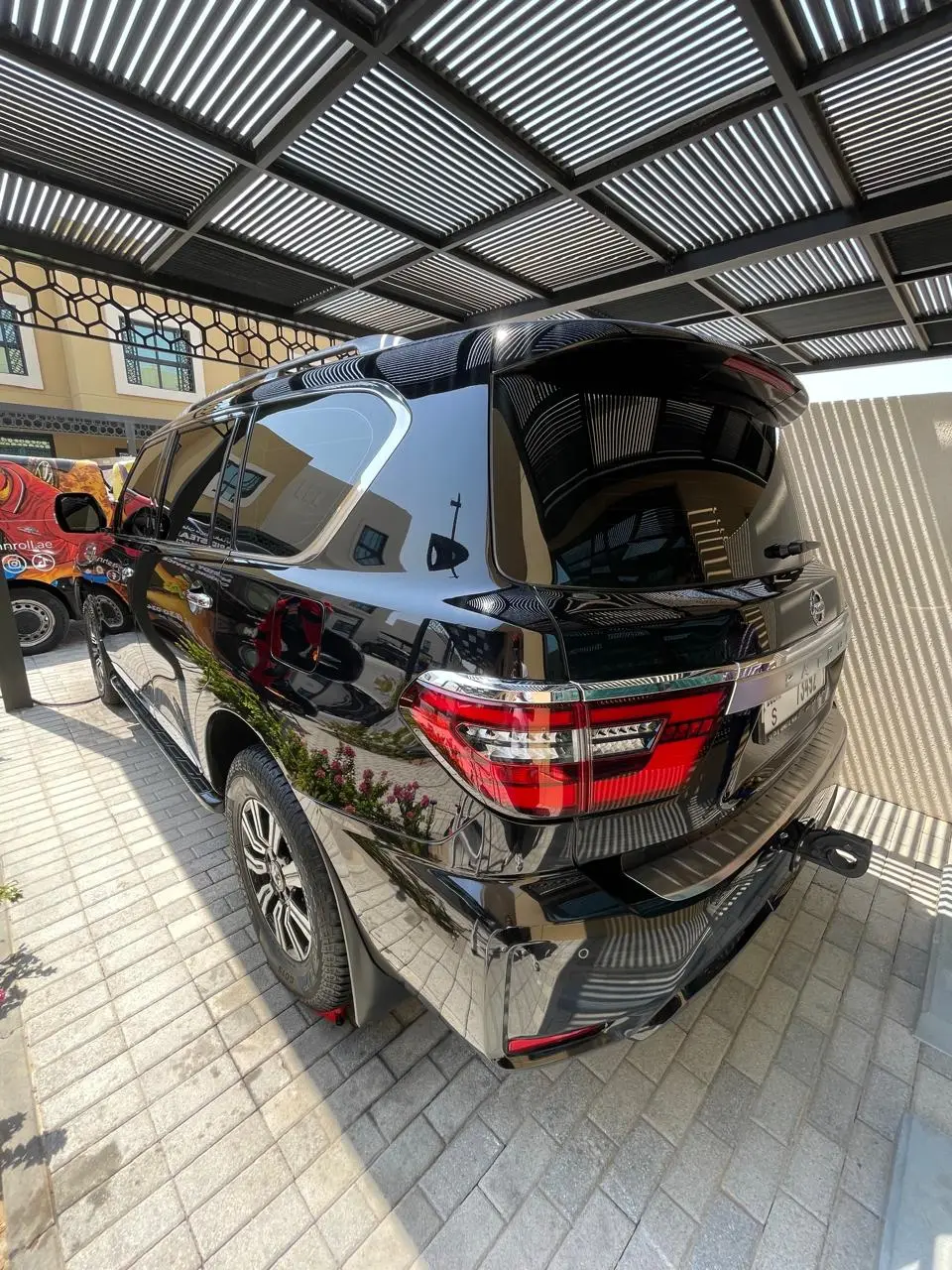 Rear and side view of a glossy black SUV parked on paving beneath a slatted metal canopy. A partially visible vehicle with colorful graphics is parked beside it.