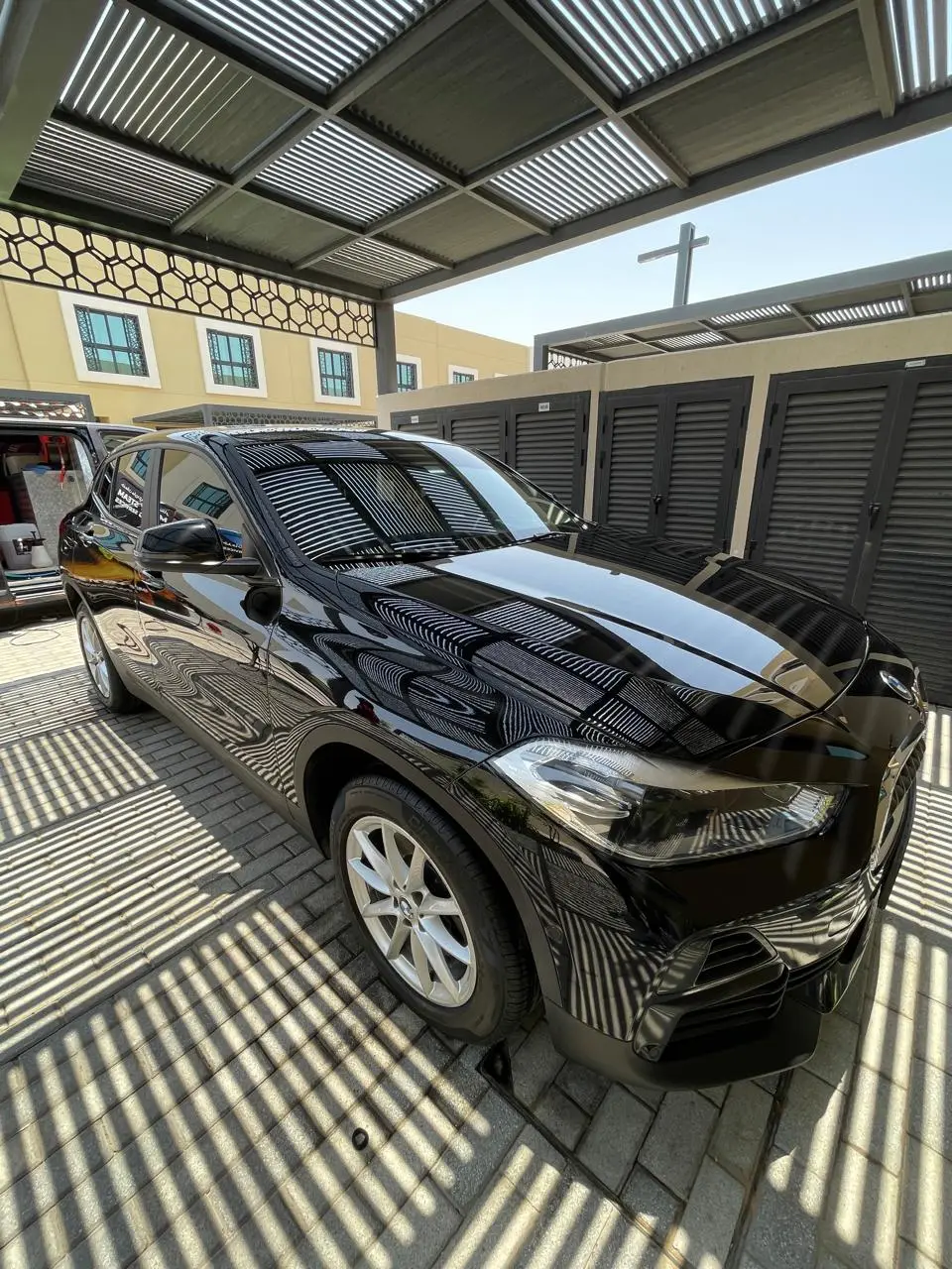 A glossy black BMW SUV parked beneath a slatted carport, with strong reflections across its hood and sides. An open van with supplies is partially visible behind it.