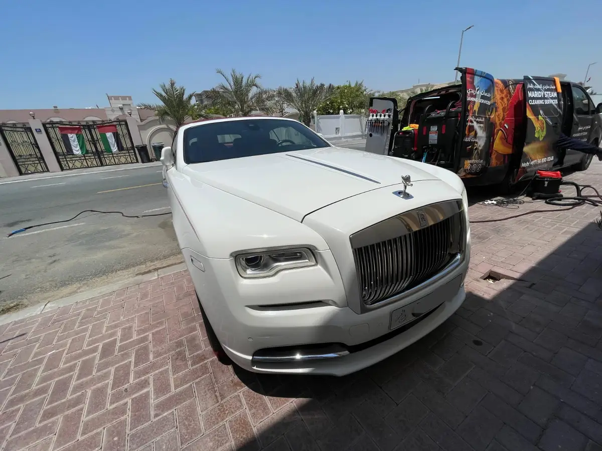 A white Rolls-Royce parked beside an open, branded steam-cleaning van with cleaning machines, bottles and hoses visible. UAE flags hang on gates across the street.