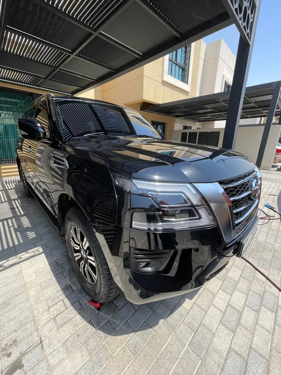 A glossy black Nissan SUV parked under a slatted metal carport beside a house, with a hose visible on the paved driveway.